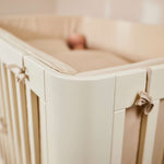 FLEXA baby bed, 140x70 cm, cream finish, visible soft padding, child resting inside; nursery context.