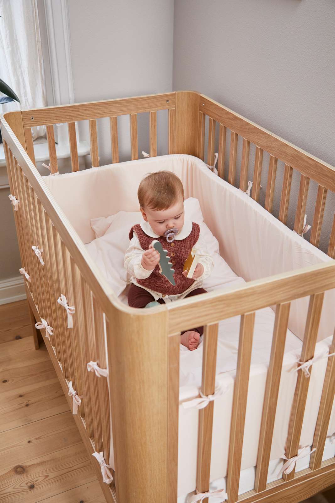 Baby with pacifier in FLEXA baby bed, OAK, soft bedding and bumpers, nursery; child-safe rails visible.