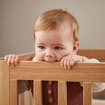 Baby in FLEXA OAK cot bed, safely biting top rail; child-safe design, oak finish, nursery setting visible.